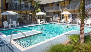 The image shows an outdoor swimming pool area with umbrellas, lounge chairs, and people enjoying the water. There are hotel balconies surrounding the pool.