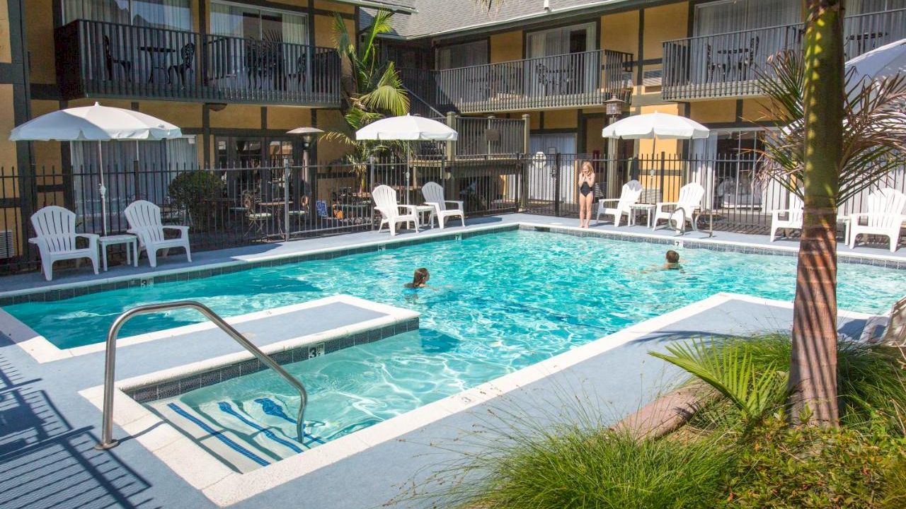 The image depicts an outdoor pool at a motel or hotel, with several people swimming and lounging, and some chairs and umbrellas surrounding the pool area.