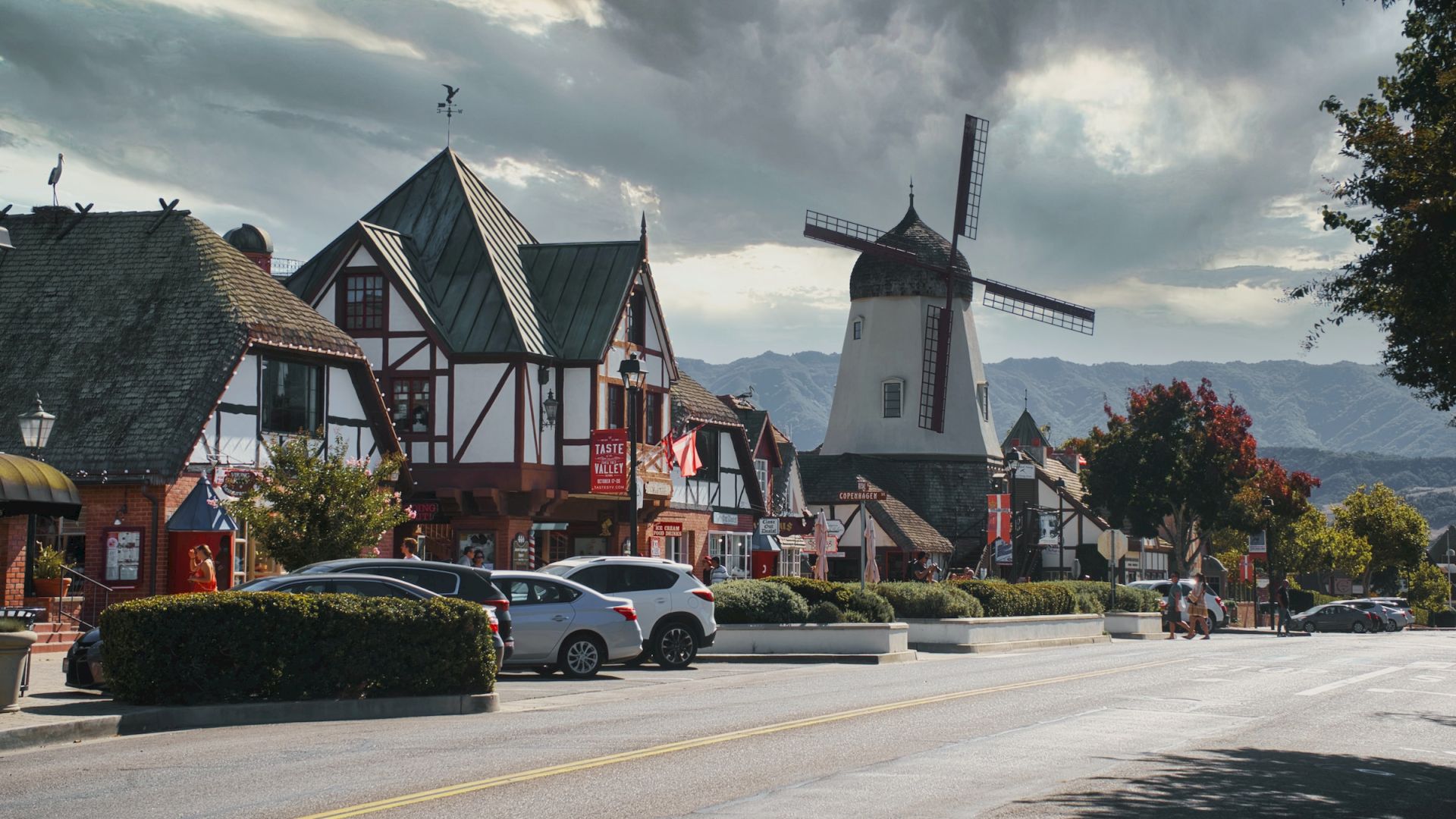 The image shows a street lined with buildings in a quaint village, featuring a prominent windmill with cloudy skies overhead.