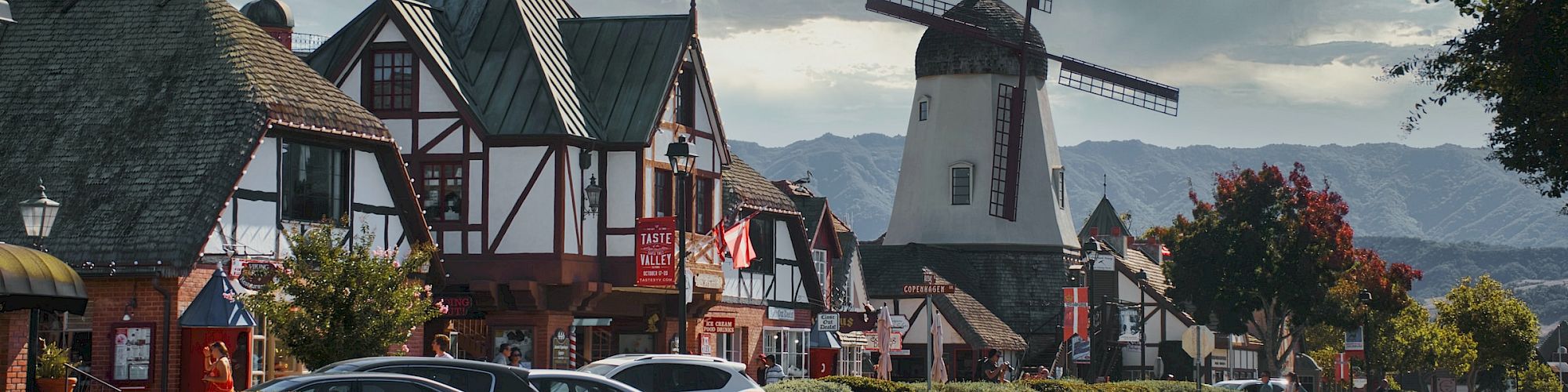 The image shows a street lined with buildings in a quaint village, featuring a prominent windmill with cloudy skies overhead.