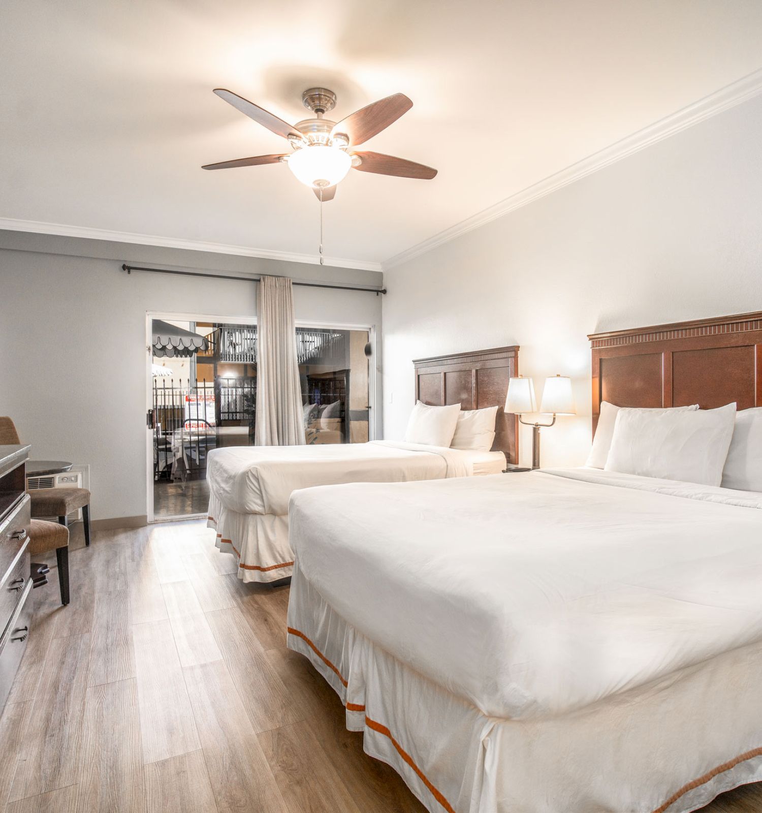 This image shows a hotel room with two large beds, a ceiling fan, a desk, chairs, and large windows allowing natural light to enter the room.