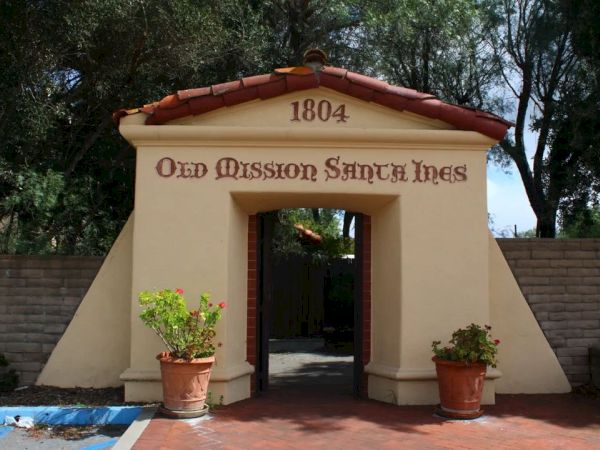 The image shows the entrance to Old Mission Santa Ines, with the year 1804 inscribed at the top and two potted plants beside the doorway.