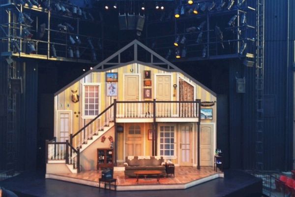 This image shows a theater stage set resembling the interior of a house, with an audience gathering in front. The set includes stairs and furniture.