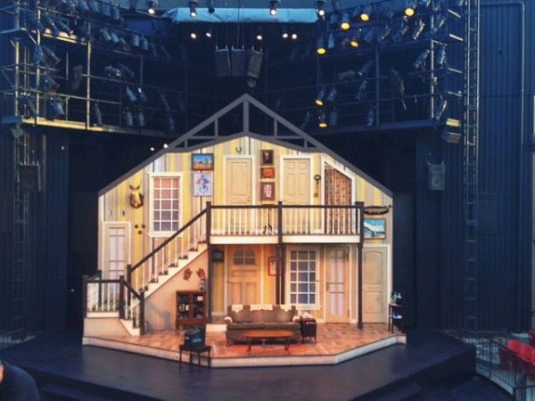 This image shows a theater stage set resembling the interior of a house, with an audience gathering in front. The set includes stairs and furniture.