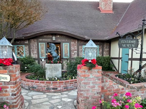 A quaint building with a pitched roof, brick path, statue, and garden. Sign reads “ELVERHØJ MUSEUM.”.