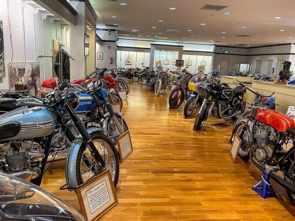 A museum room filled with a variety of vintage motorcycles displayed on a polished wooden floor with informational plaques.