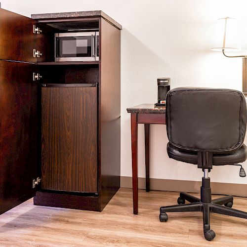 A small workspace with a desk, chair, lamp, coffee maker, microwave, and a TV cabinet, possibly in a hotel room.