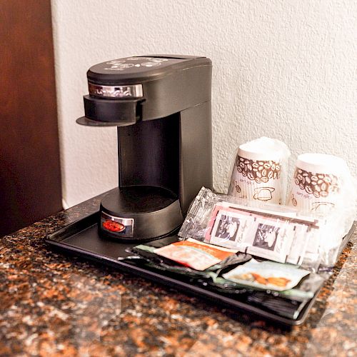 A single-serve coffee maker is displayed on a countertop, accompanied by paper cups and coffee condiments organized on a tray.