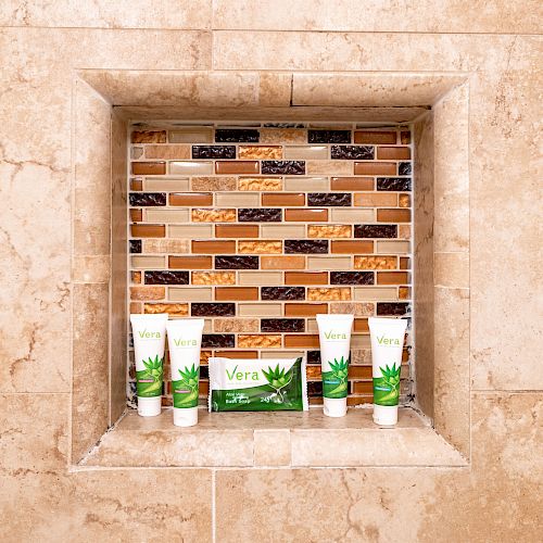 The image shows a shower niche with aloe vera products, including tubes and a soap bar, set against a decorative mosaic tile backdrop.