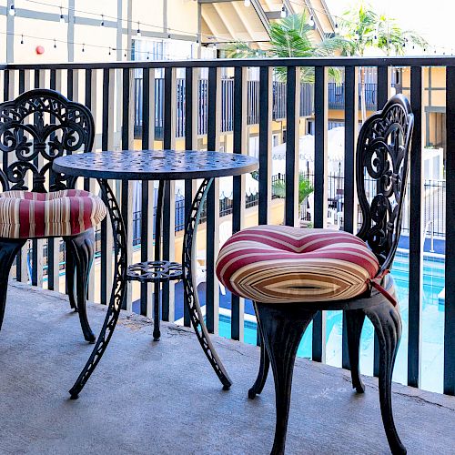 A small balcony with two wrought-iron chairs, a round table, striped cushions, and a view of a pool area surrounded by a railing ends the sentence.