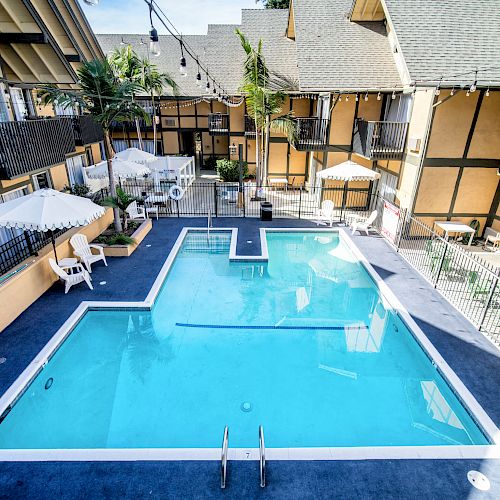 An outdoor swimming pool area at a hotel, surrounded by lounge chairs, umbrellas, and tan buildings with balconies.