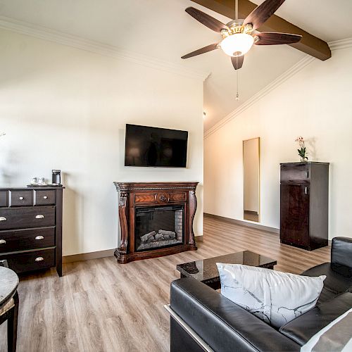 A living room with a black sofa, wooden furniture, wall-mounted TV, fireplace, ceiling fan, and decorative items, including flowers and framed art.