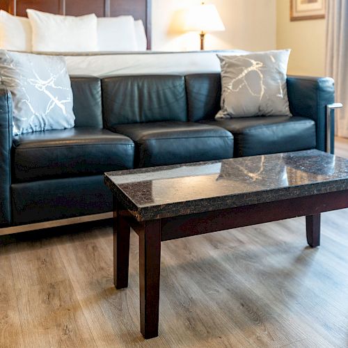 A modern living space with a black leather sofa, patterned pillows, a wooden coffee table, and part of a bed and chair in the background.