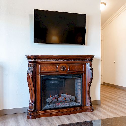 The image showcases a room with a wall-mounted TV above a wooden electric fireplace, a mirror, and a portion of a dark dresser.