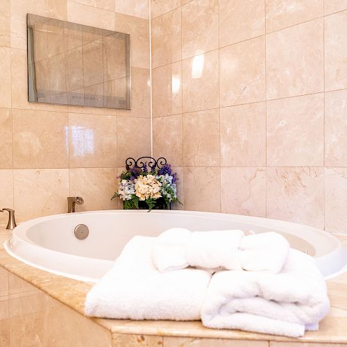 A luxurious bathroom features a large bathtub, folded towels, toiletries, and a decorative floral arrangement, creating an elegant and relaxing space.