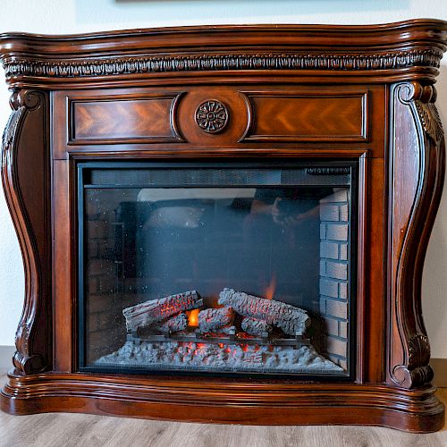 An ornate wooden electric fireplace with a glass front, featuring realistic looking logs and flames, stands against a wall on a wooden floor.