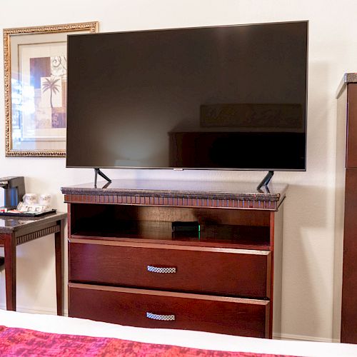 The image shows a room setup with a TV on a cabinet, a desk with a coffee maker and chair, a picture frame, and an orchid on the desk.