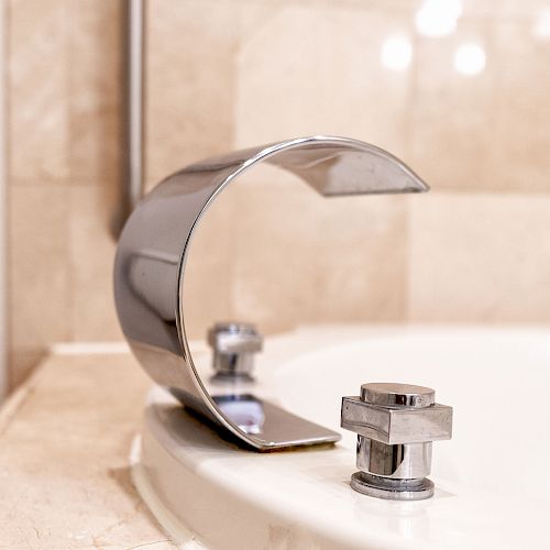 This image shows a close-up of a modern, chrome-plated faucet on the edge of a bathtub, with beige tiles in the background.