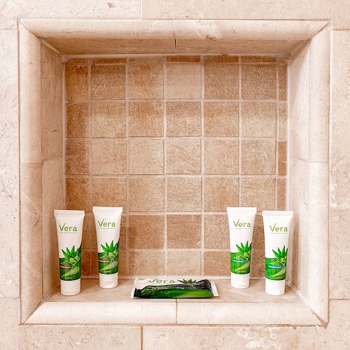 A tiled shower niche holding five skincare products, including tubes and a small packet, all labeled with "Vera" and featuring images of aloe leaves.