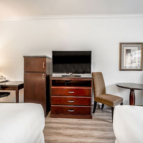 A hotel room with two beds, a TV on a dresser, a mini-fridge, a desk with a chair and lamp, and a small table with chairs.