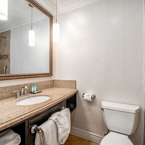 A modern bathroom features a large mirror, sink with granite countertop, toilet, and a shower area with beige tiles and a handrail.