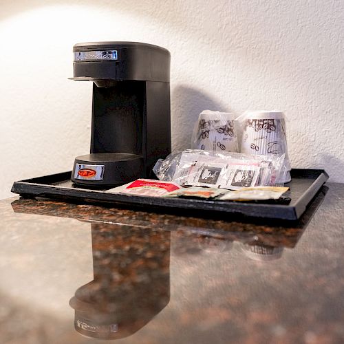 A coffee maker on a tray with disposable cups, coffee packets, sugar, and stir sticks is placed on a countertop.