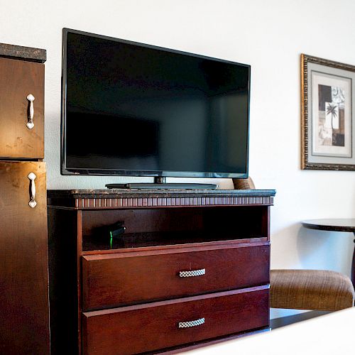 A hotel room features a TV on a wooden TV stand, a fridge, a framed picture, and a small table with two chairs.