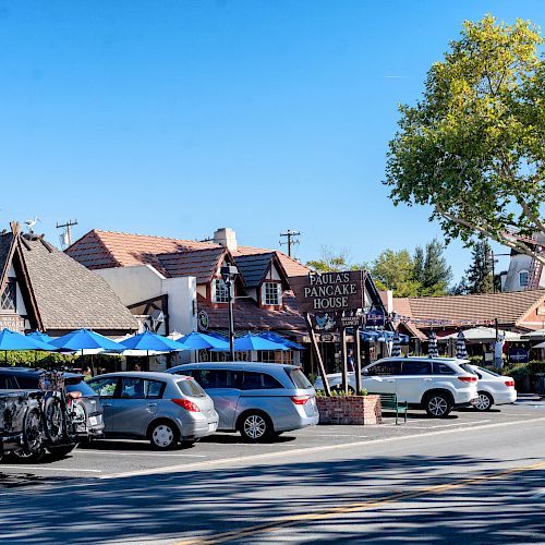 The image shows a quaint street with parked cars, outdoor dining, and businesses including Paula's Pancake House, under a clear blue sky.