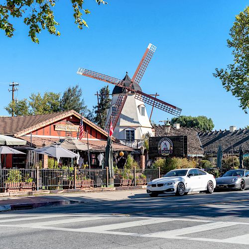 A street view featuring a building with a windmill, some parked cars, a pedestrian crossing, and outdoor seating under umbrellas.