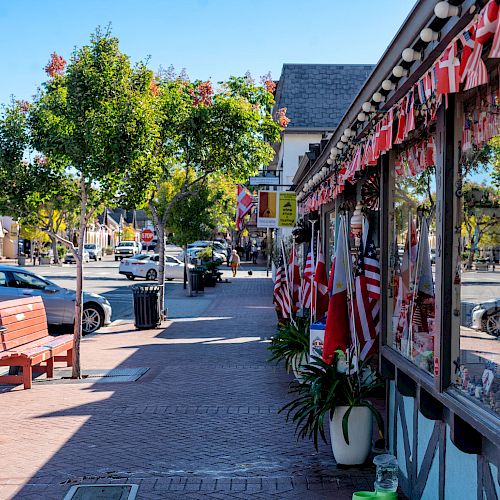 A quaint street with shops decorated with flags, a few parked cars, benches, trees, and a clear blue sky. The setting is bright and inviting.