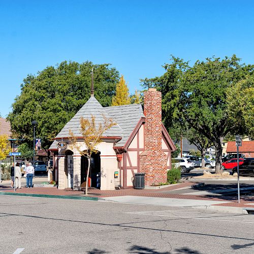 A small, quaint building with Tudor-style architecture sits on a street corner, surrounded by trees, cars, and a few people standing nearby.