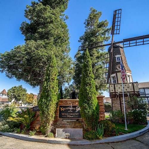 The image shows a quaint entrance with a sign for "Korshamps Inn" and a windmill structure, surrounded by lush greenery and a clear blue sky.