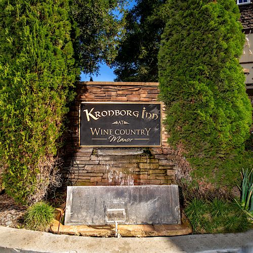 A sign for Krondborg Inn, titled "AUTH. Wine Country Manor," is nestled between trees with plants around, set in a landscaped area.