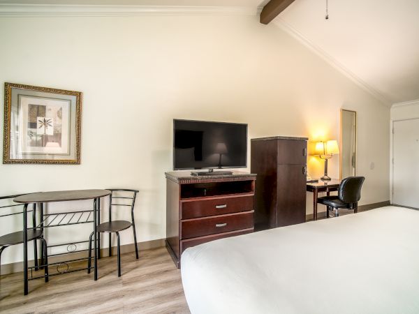 A hotel room with a TV, dresser, small dining table, framed artwork, desk with lamp, and bed. The room has wooden flooring and white walls.