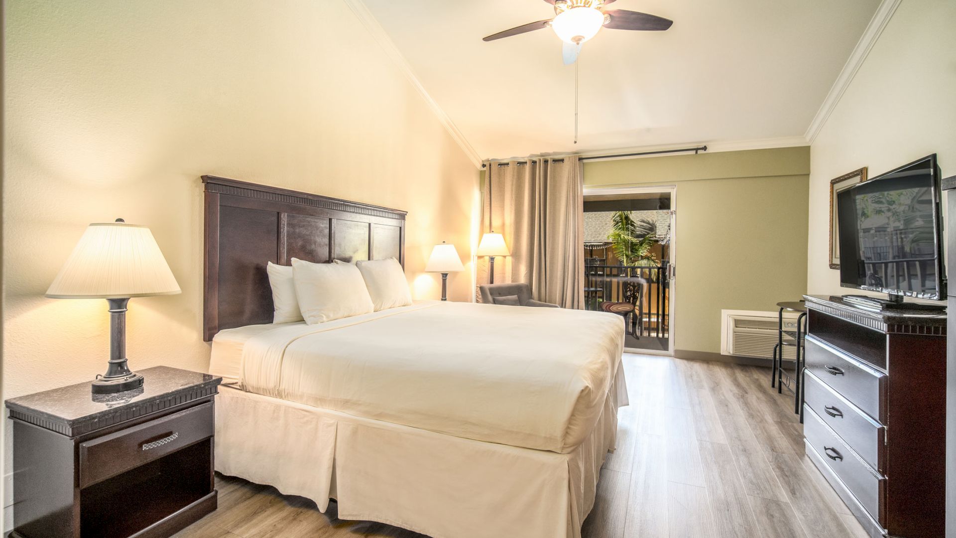 A neatly arranged hotel room with a king-sized bed, bedside tables, lamps, a ceiling fan, a dresser with a TV, and a balcony area in the background.
