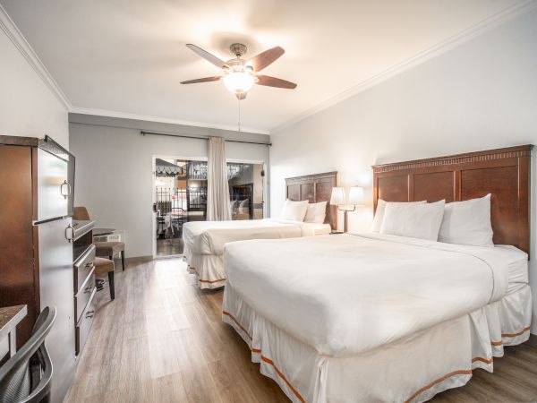 A hotel room with two queen beds, a ceiling fan, desk, chairs, and a dresser. The room is well-lit with modern decor and wooden flooring.