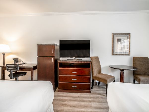 A hotel room features two beds, a desk with a lamp, a TV on a dresser, a mini fridge, a chair, and a small table with two chairs.