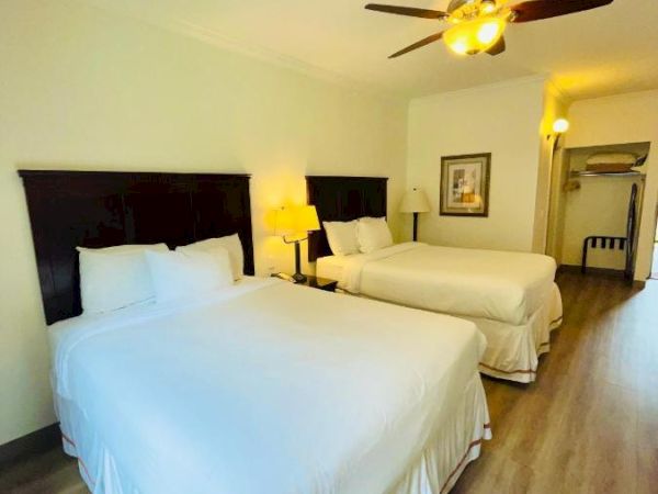 The image shows a hotel room with two neatly made double beds, a ceiling fan, a bedside lamp, and a framed picture on the wall.
