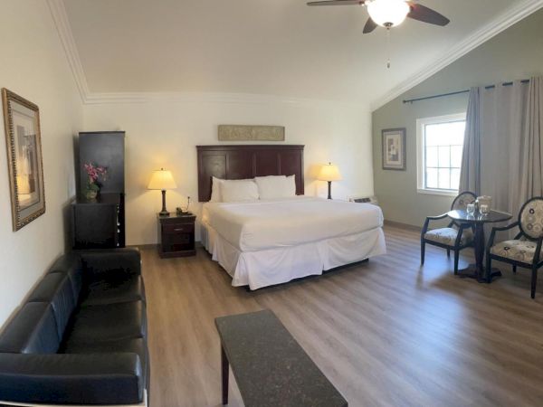 A spacious hotel room with a large bed, sofa, table, chairs, nightstands, lamps, artwork, a ceiling fan, and a window with curtains ending the sentence.