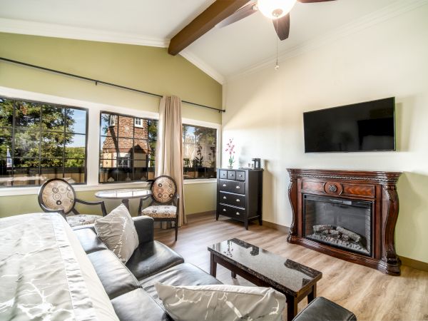A cozy living room with a couch, coffee table, TV, fireplace, and dresser. Large windows and a ceiling fan complete the space.