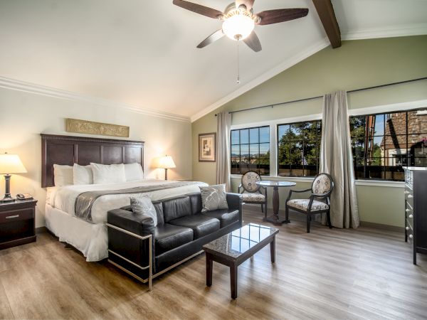 A spacious, well-lit bedroom with a bed, sofa, coffee table, and small dining set. Ceiling fan, wooden floors, and large window with curtains.