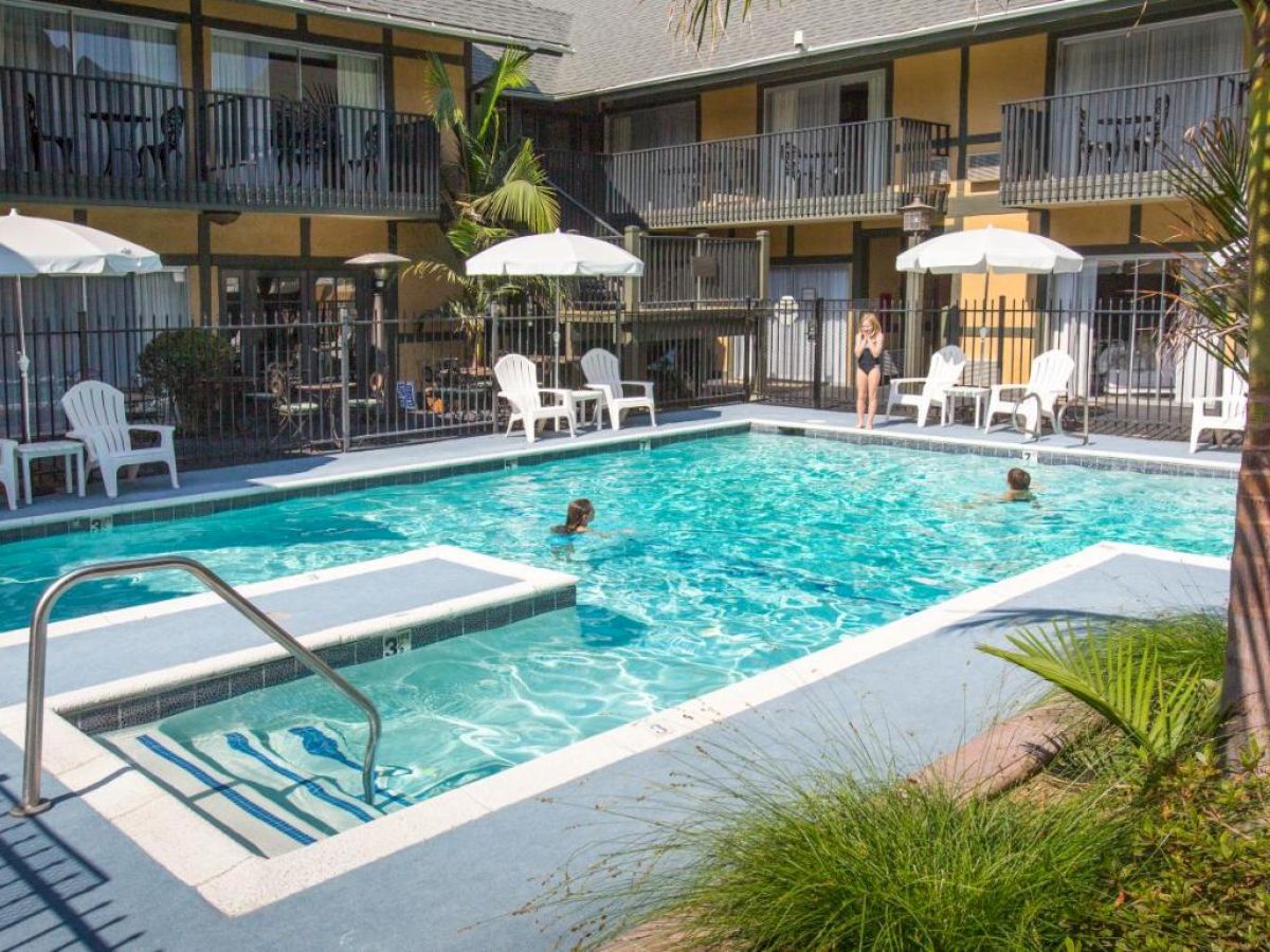 A sunny outdoor pool area with white umbrellas, lounge chairs, and palm trees surrounded by apartment-style buildings and people in the pool.