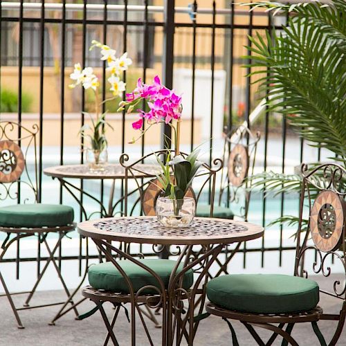 An outdoor seating area with ornate chairs and tables, topped with floral centerpieces and surrounded by greenery, near a pool.