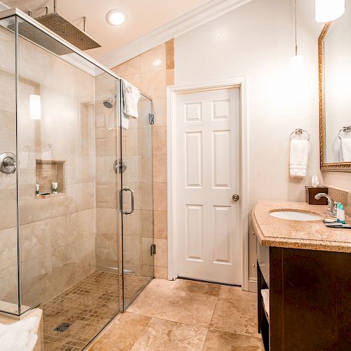 The image shows a modern bathroom with a glass shower enclosure, a vanity with a sink, a large mirror, and beige tile flooring and walls.