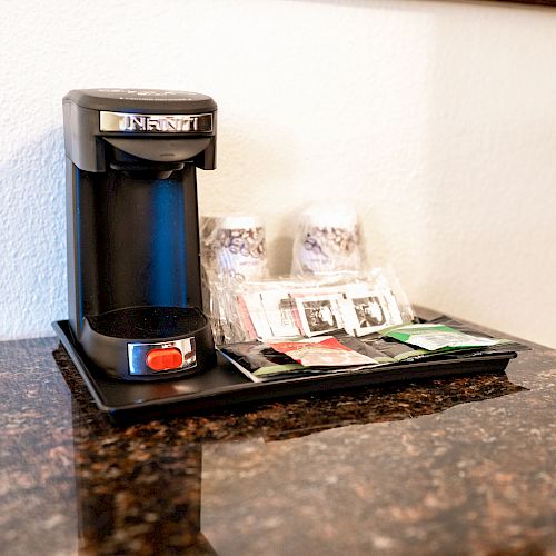 The image shows a single-serve coffee maker on a counter with coffee packets, condiments, and disposable cups on a tray next to it.