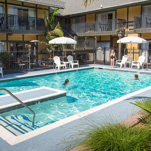 An outdoor pool area with lounge chairs, umbrellas, and a small jacuzzi. A few people are swimming, and there are plants near the poolside.