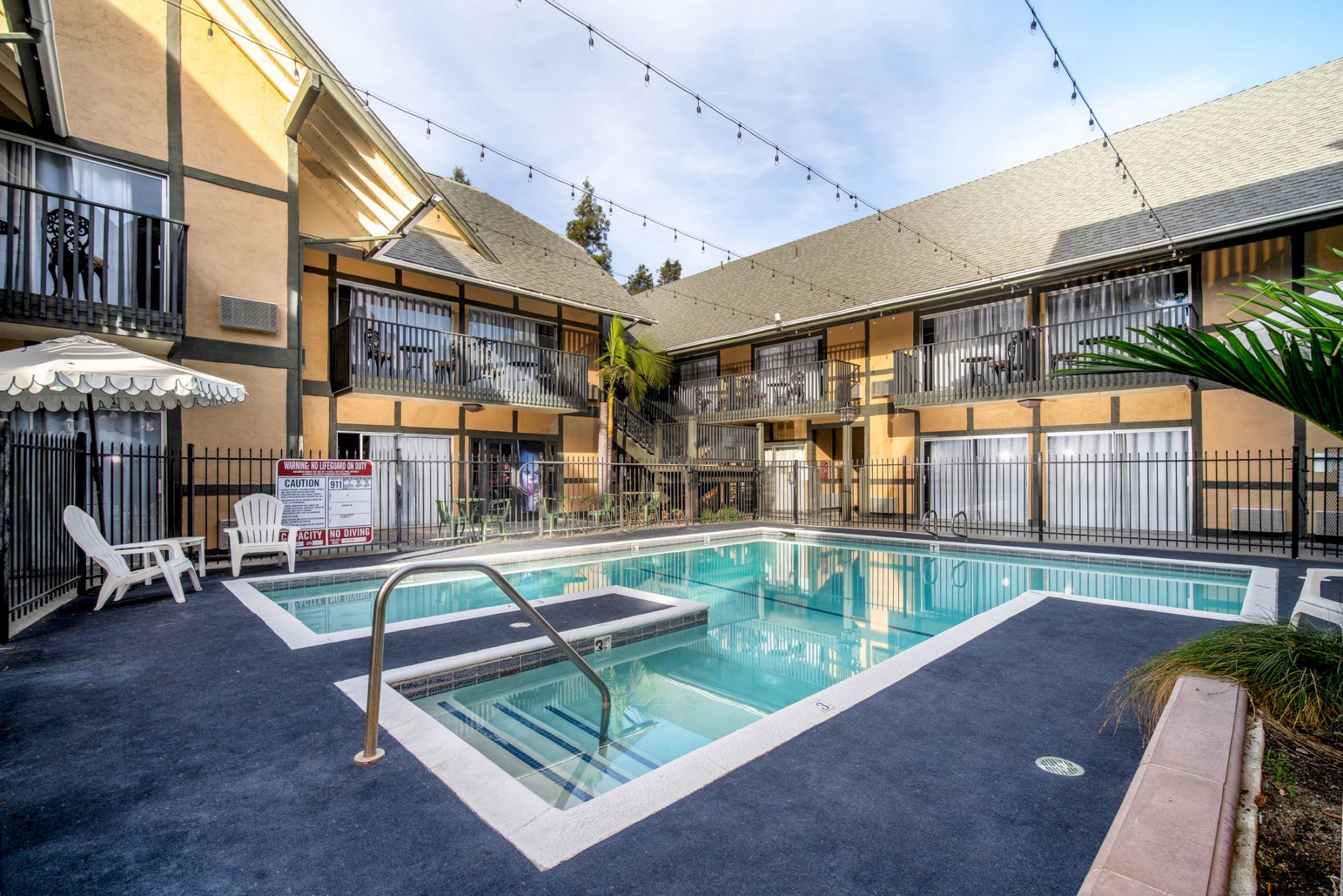 An outdoor courtyard with a swimming pool, lounge chairs, and string lights, surrounded by a two-story building with balconies.