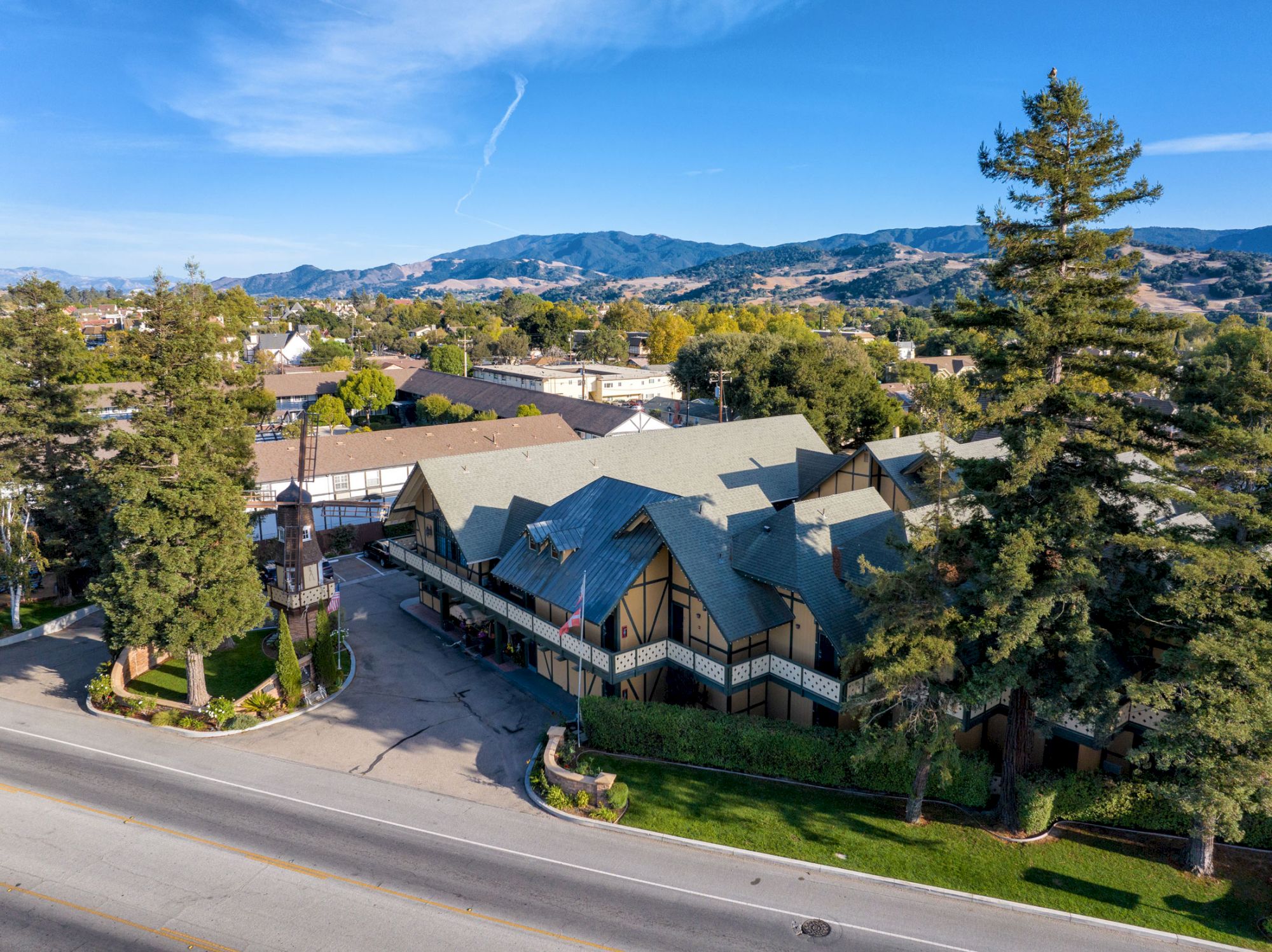 An aerial view of a building with a distinctive architectural style, surrounded by trees and a well-maintained road, with mountains in the background.