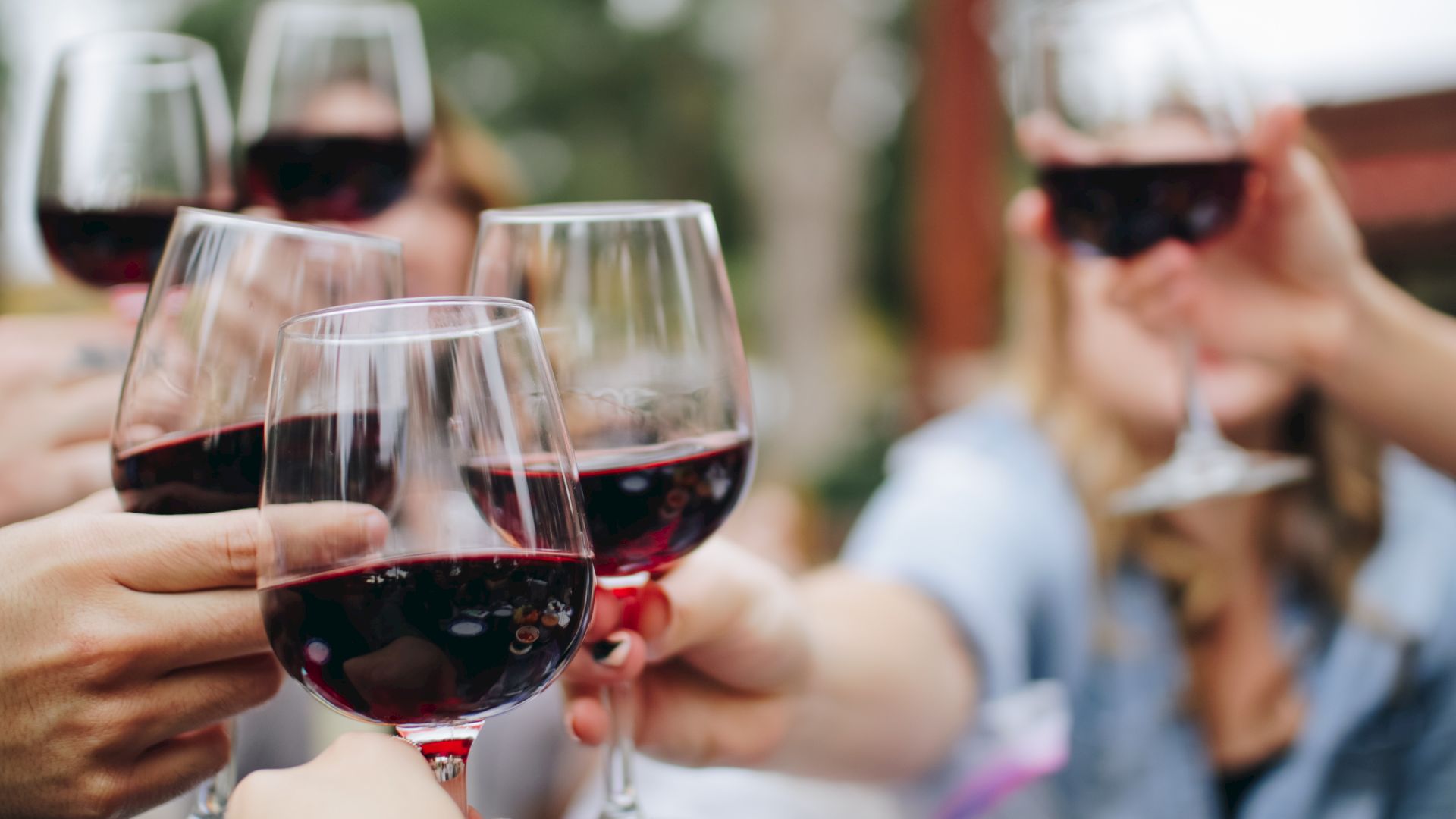 A group of people clinking glasses of red wine together in a celebratory toast, with blurred background of an outdoor setting.